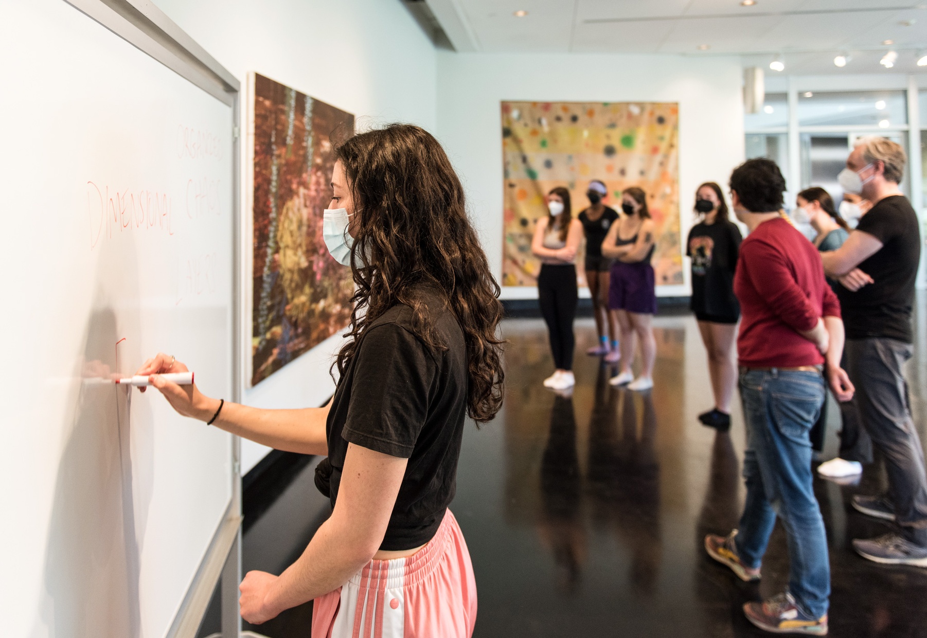A masked young woman writes on a white board while a group of people look at an abstract painting behind her.
