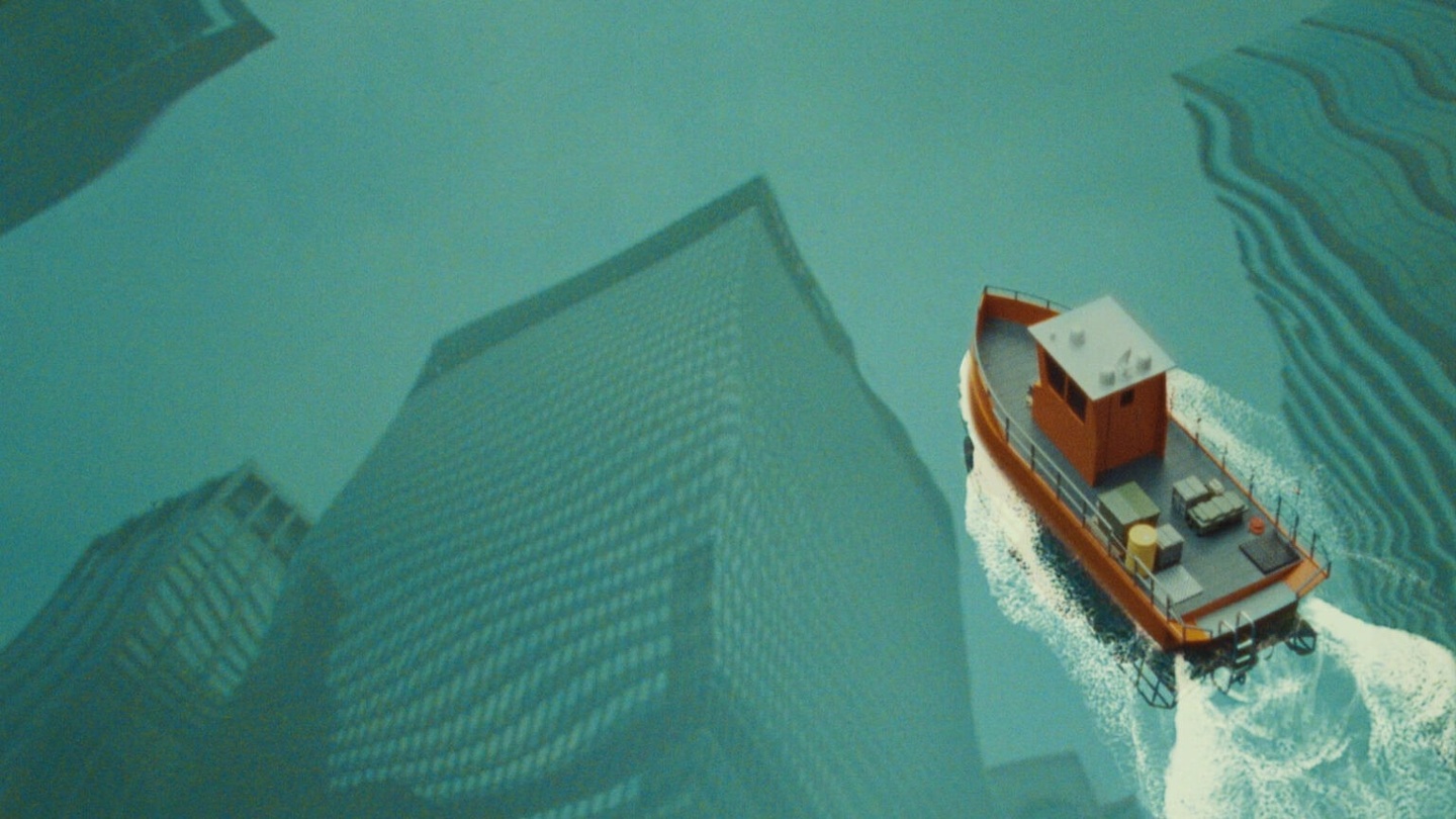 still from a film of a view from above of small boat moving across the water with reflections of skyscrapers