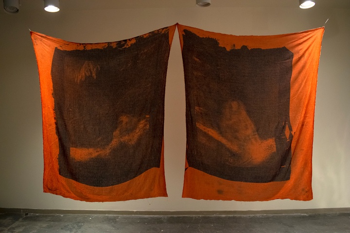 Two large orange fabric with faded black ink prints hanging side by side in a gallery space