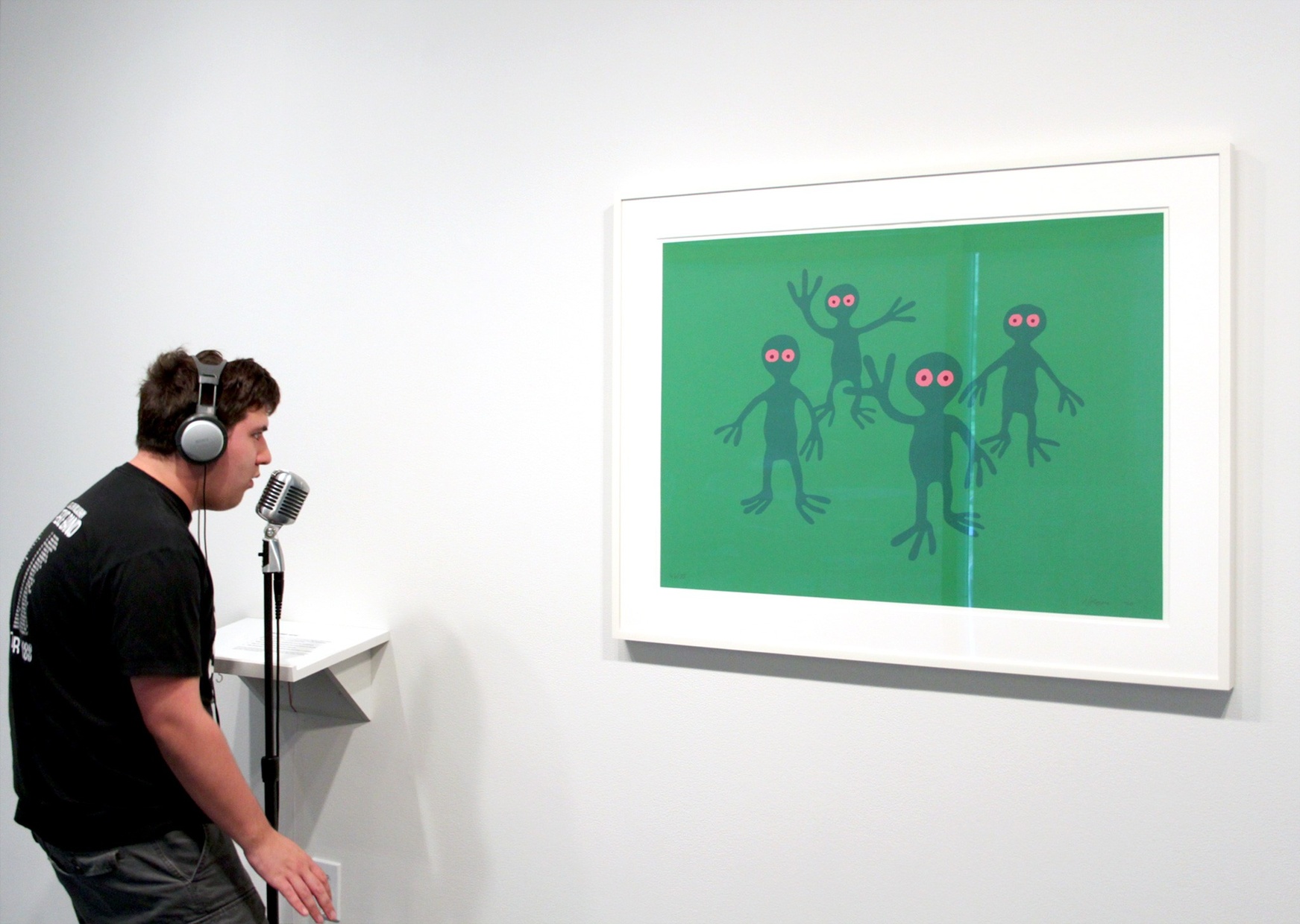 A young, light-skinned man wearing big, silver headphones speaks into a microphone while facing an abstract, green painting of four aliens hung on a white wall.