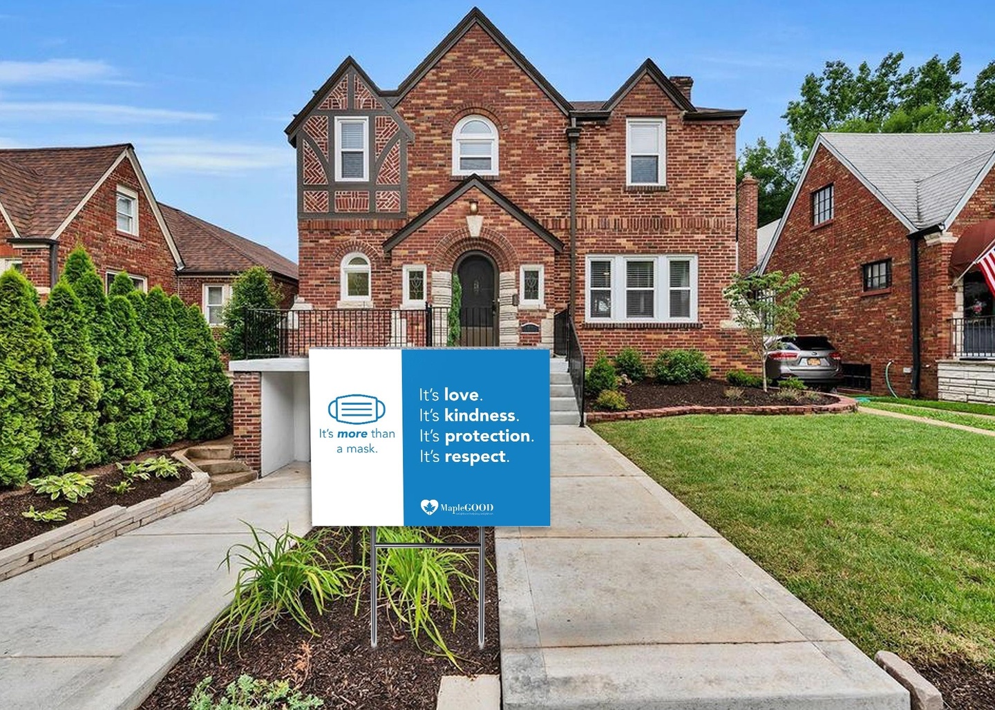 Blue-and-white yard sign advocating for people to wear masks to support public health, displayed in a narrow planting bed outside a two-story brick home.