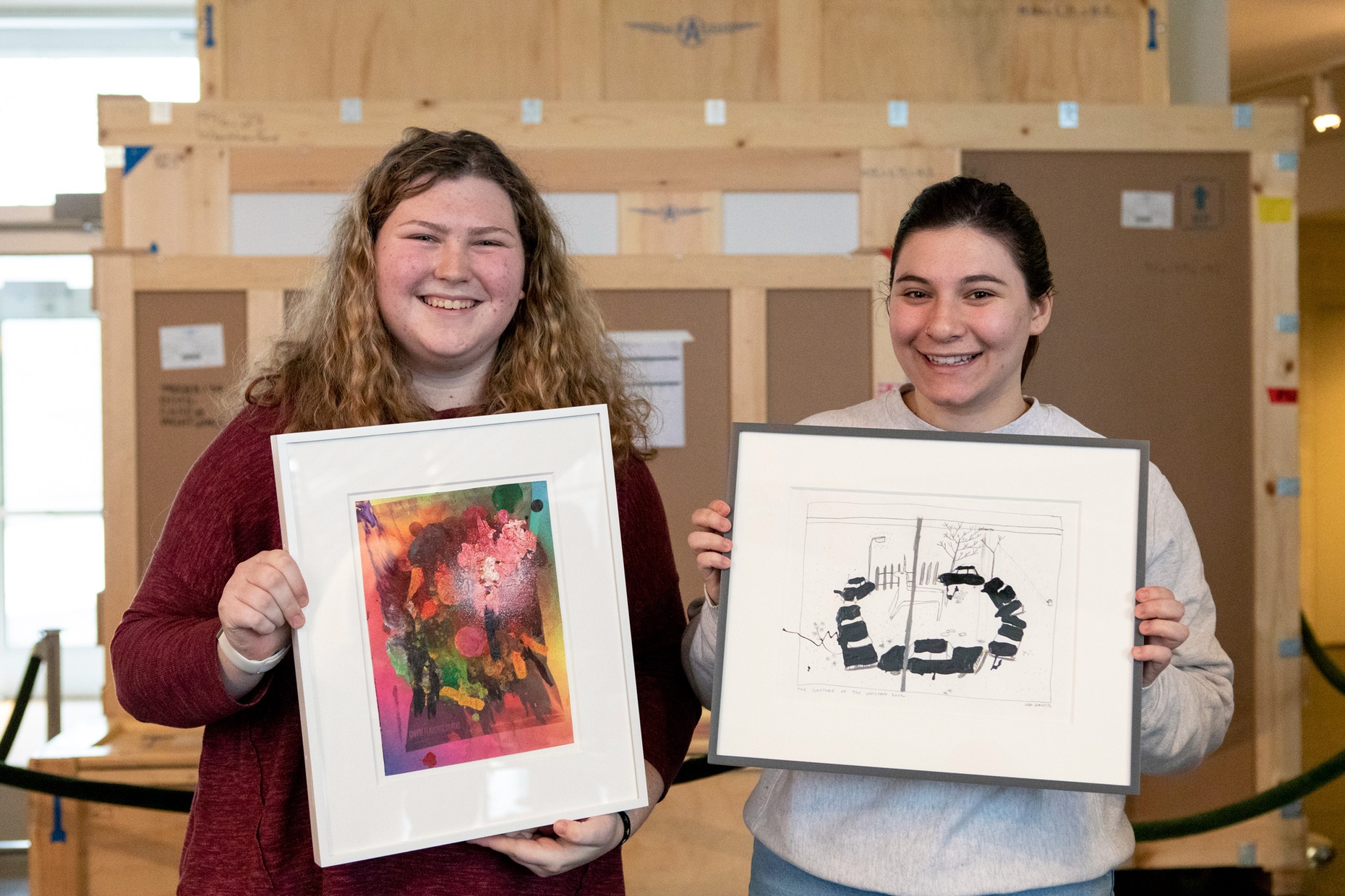 Two smiling students each hold a framed artwork in front of them, large wooden shipping crates are in the background.