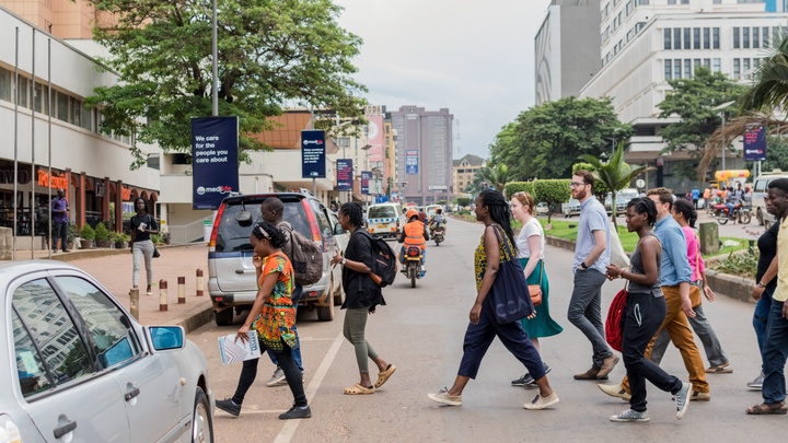 Group of people crossing a street in Kampala, Uganda. Multi-story apartment and office buildings can be seen down the road, and several people on mopeds ride by.