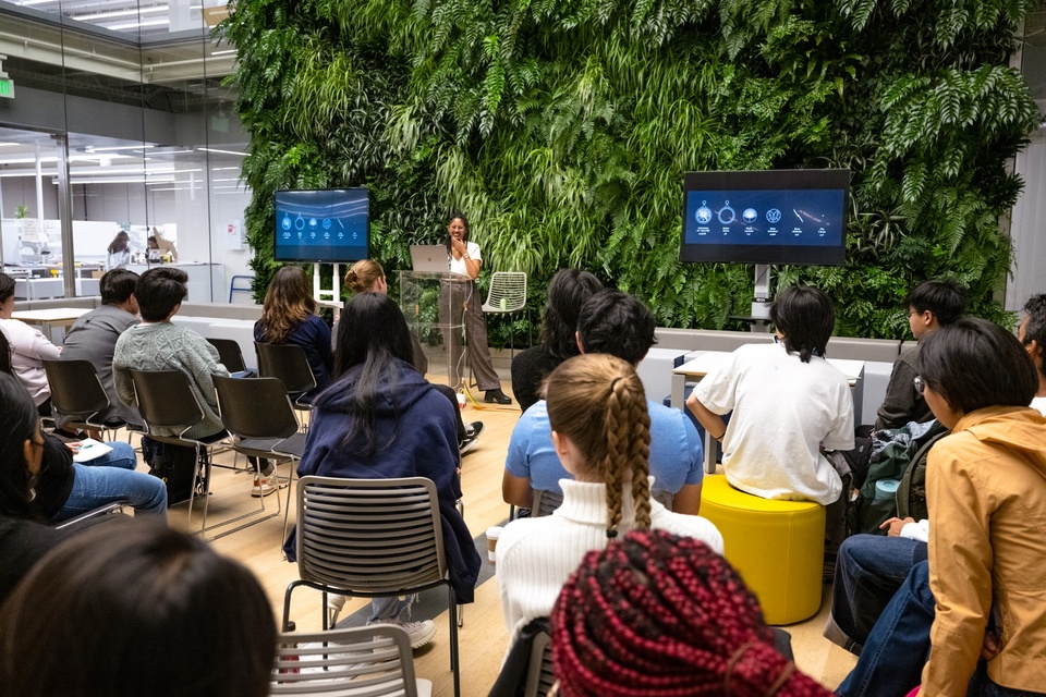 Person delivers a lecture to a room of students. Behind them is a living green wall and two video screens with video game iconography on them.