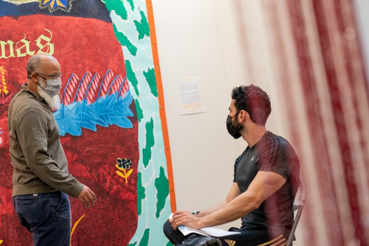 Low angle shot of a person standing next to a large scale fabric wall hanging, conversing with another person seated in front of it.
