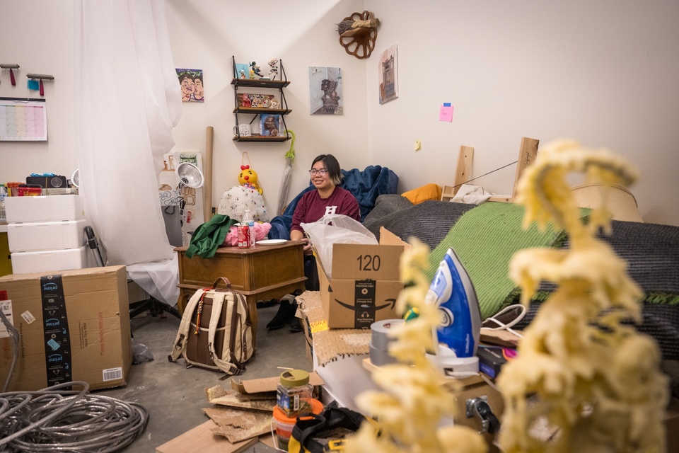 Student sits in an armchair in the back corner of a studio space full of sculptural work, cardboard, fabric, storage bins, duct tape, an iron, and various knick-knacks on a shelf.