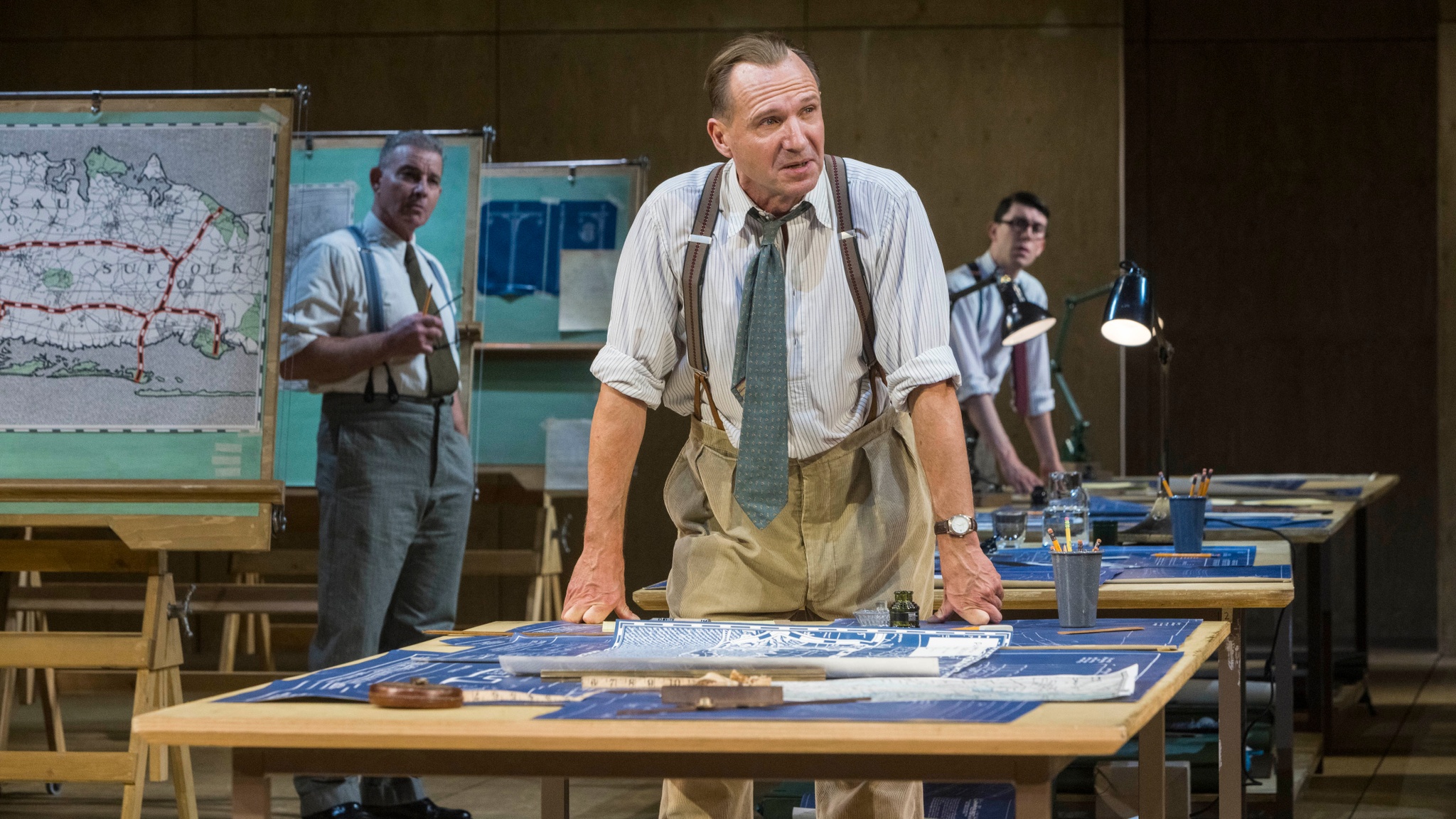 The actor Ralph Fiennes, a white man, stands leaning with both hands on a draftsman's table. He is in costume as Robert Moses, wearing khaki pants, suspenders, and a blue gray blue tie. In the background are two other actors dressed similarly looking toward him.
