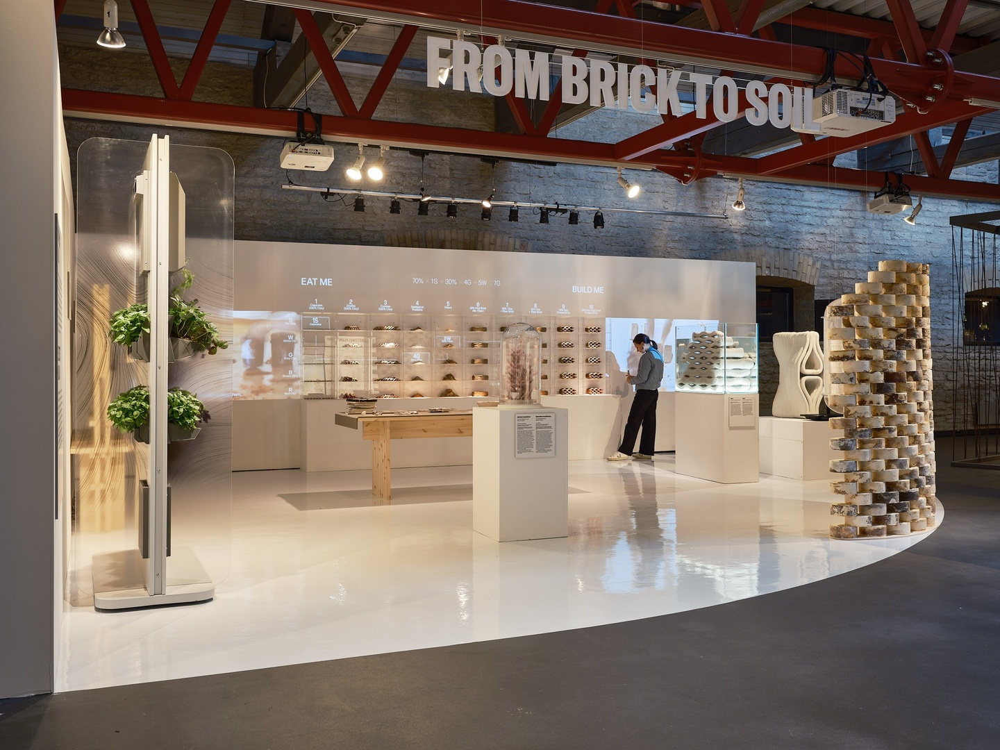 photo of a white interior space with a display of shelves titled "From Brick to Soil". A woman looks at the display 