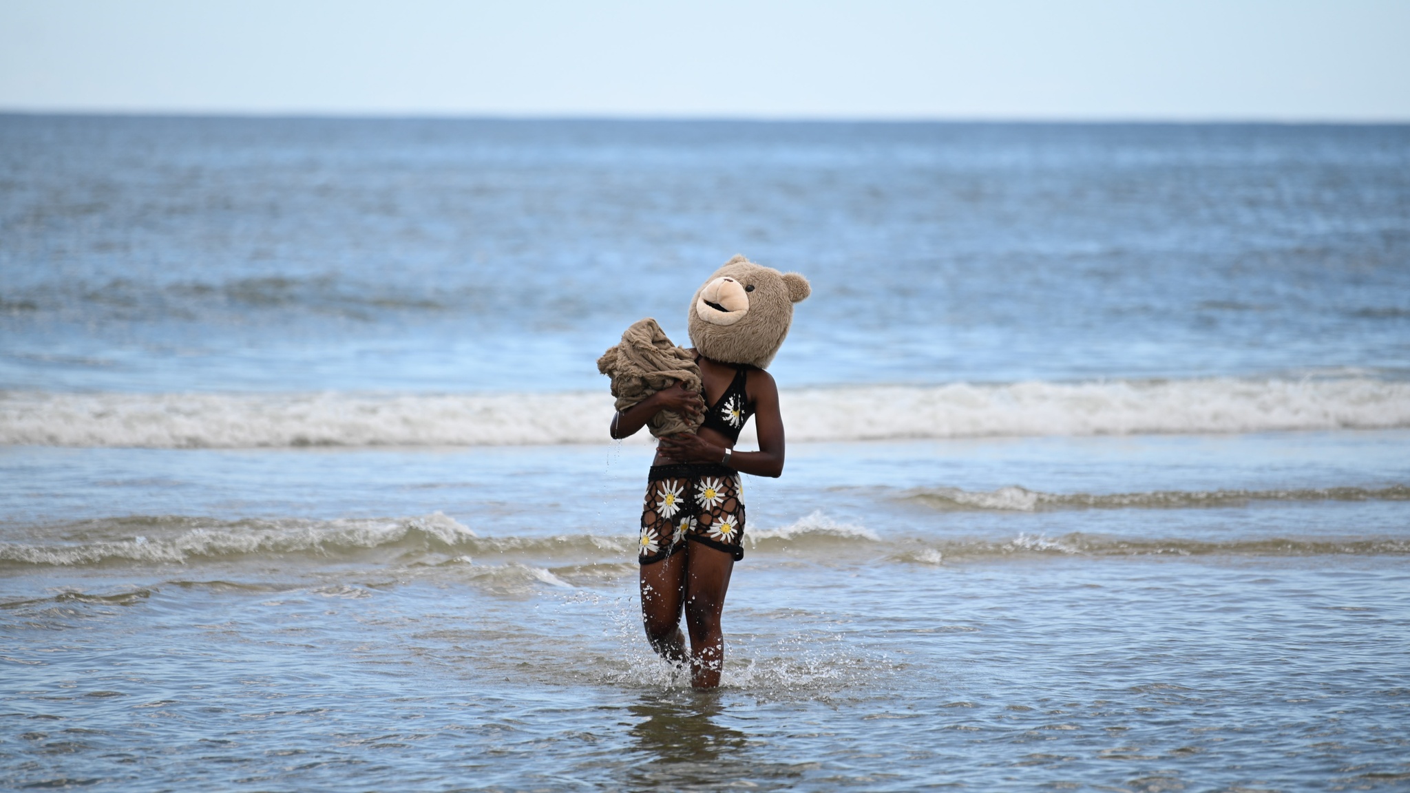 A Black person wearing a crocheted two-piece bathing suit with a yellow daisy design and a brown teddy bear costume head wades in the shallows of an ocean along a beach. They carry a bundle of brown fabric swaddled in their arms.