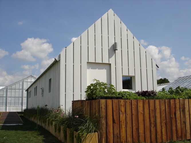 Front view of single-story gabled house with vertical white slatted siding and tall garden boxes surrounding.