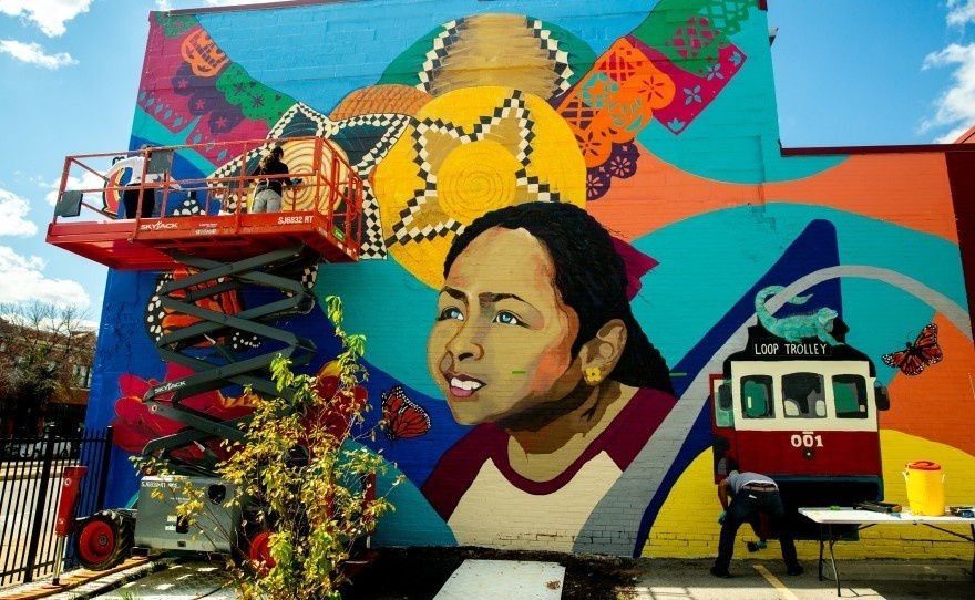 Colourful mural painting featuring a young girl gazing towards blue skies. Surrounding her are flowers, a papel picado, African baskets and orange monarchs