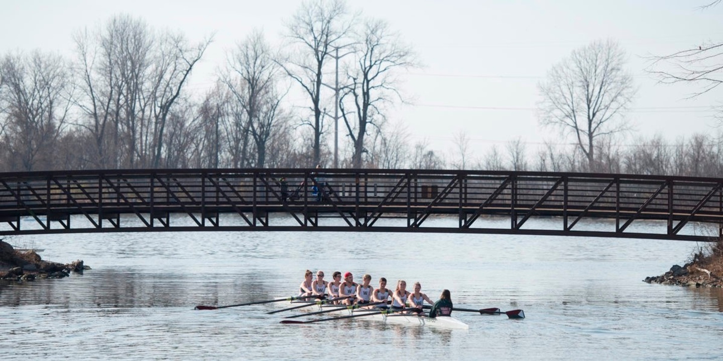 A crew team rows under a bridge on a river in the winter