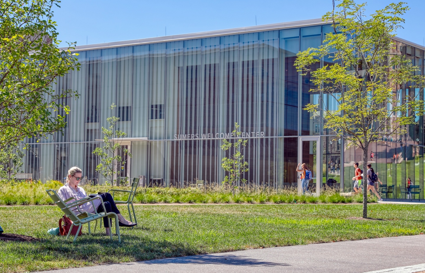 Person sits in a metal lawn chair in the shade of a little tree in front of a glass-walled building labeled Sumers Welcome Center.