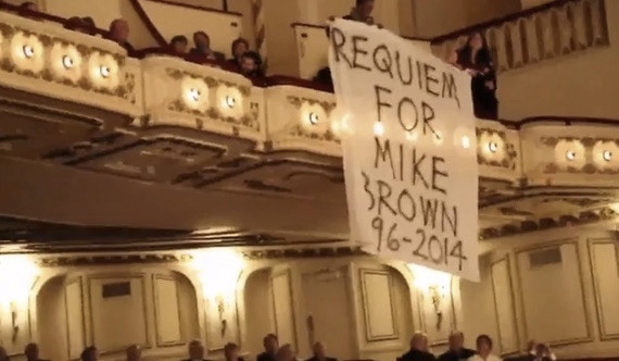 Interior of a concert hall, with people in the balcony holding a banner reading Requiem for Mike Brown 96-2014.