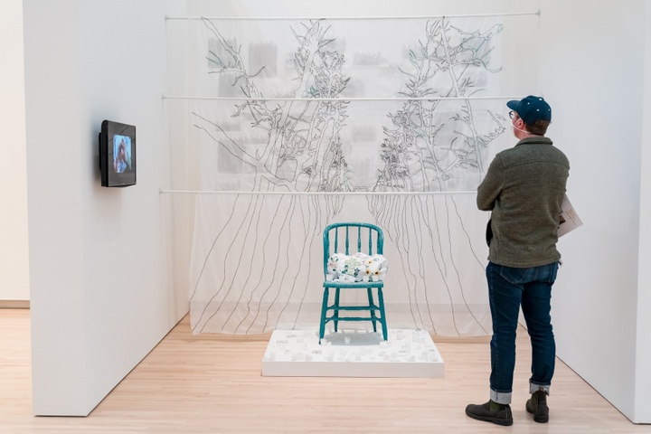 Person stands in an alcove in a gallery. Inside are hung three filmy sheets of fabric with black embroidery tracery across them. In front, on a small dias, sits a teal wooden kitchen chair set with three white pillows embroidered with flowers. Around the base of the chair are many tea candles. A video screen is affixed to the wall of the alcove but not in focus.