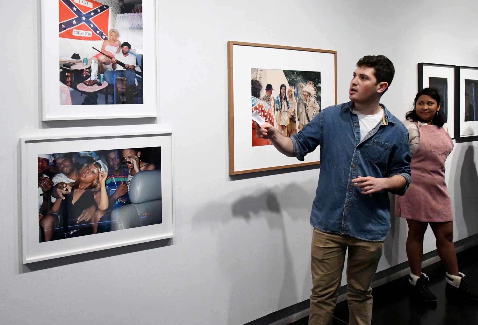A light-skinned male student in a blue button down gestures to his right at framed photographs on the wall as a female student in a pink dress looks on from behind him. 