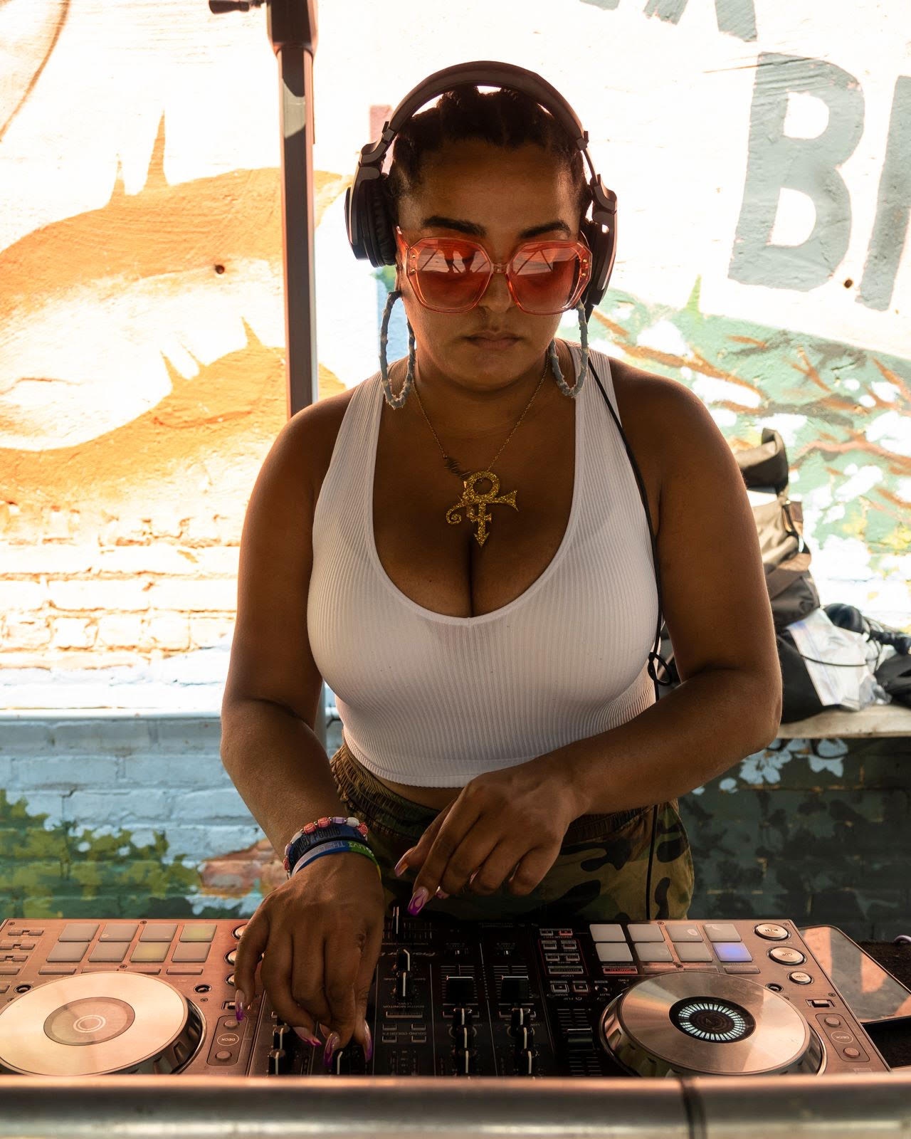 A Black woman DJs at an electronic controller. She looks down at her hands as they adjust controls on the panel. She wears a whit tank top, a gold necklace with the symbol for the Artist formerly known as Prince, and headphones. 