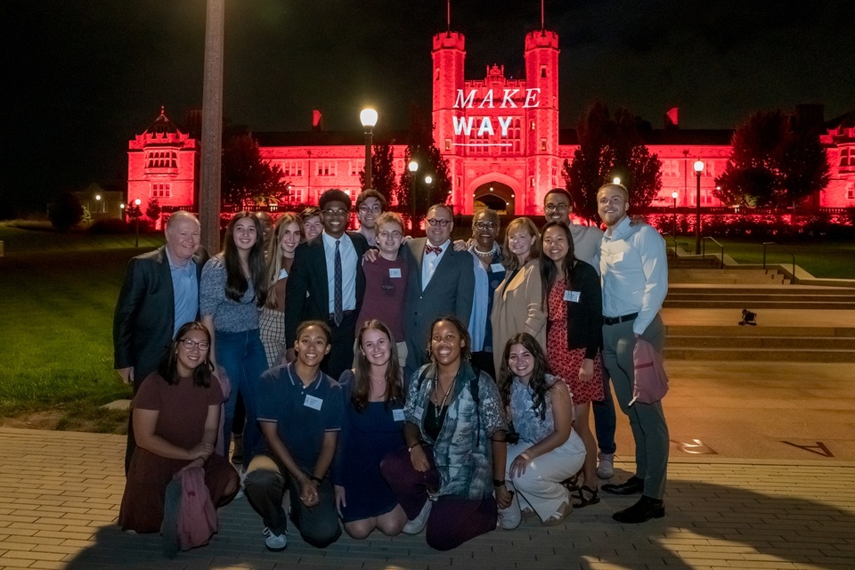 Group of students and university leadership posing together on plaza in front of Brookings at night. The building behind them is lit up with red lights and projected text reading "Make Way."