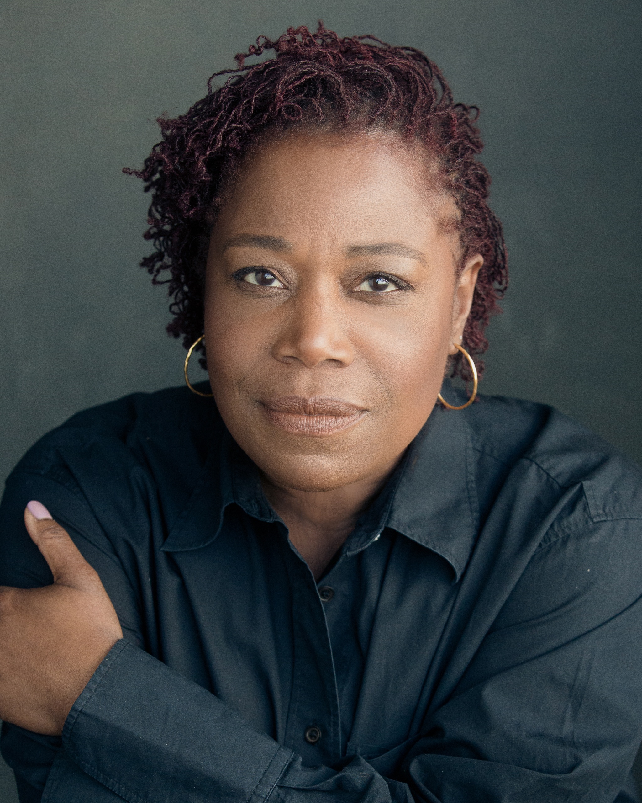 A portrait of Michele Austin, a Black actor with short auburn hair twisted in short locs and swept to the side. She wears a gray button down shirt and holds one hand up to her shoulder while looking at us with a slight smile.