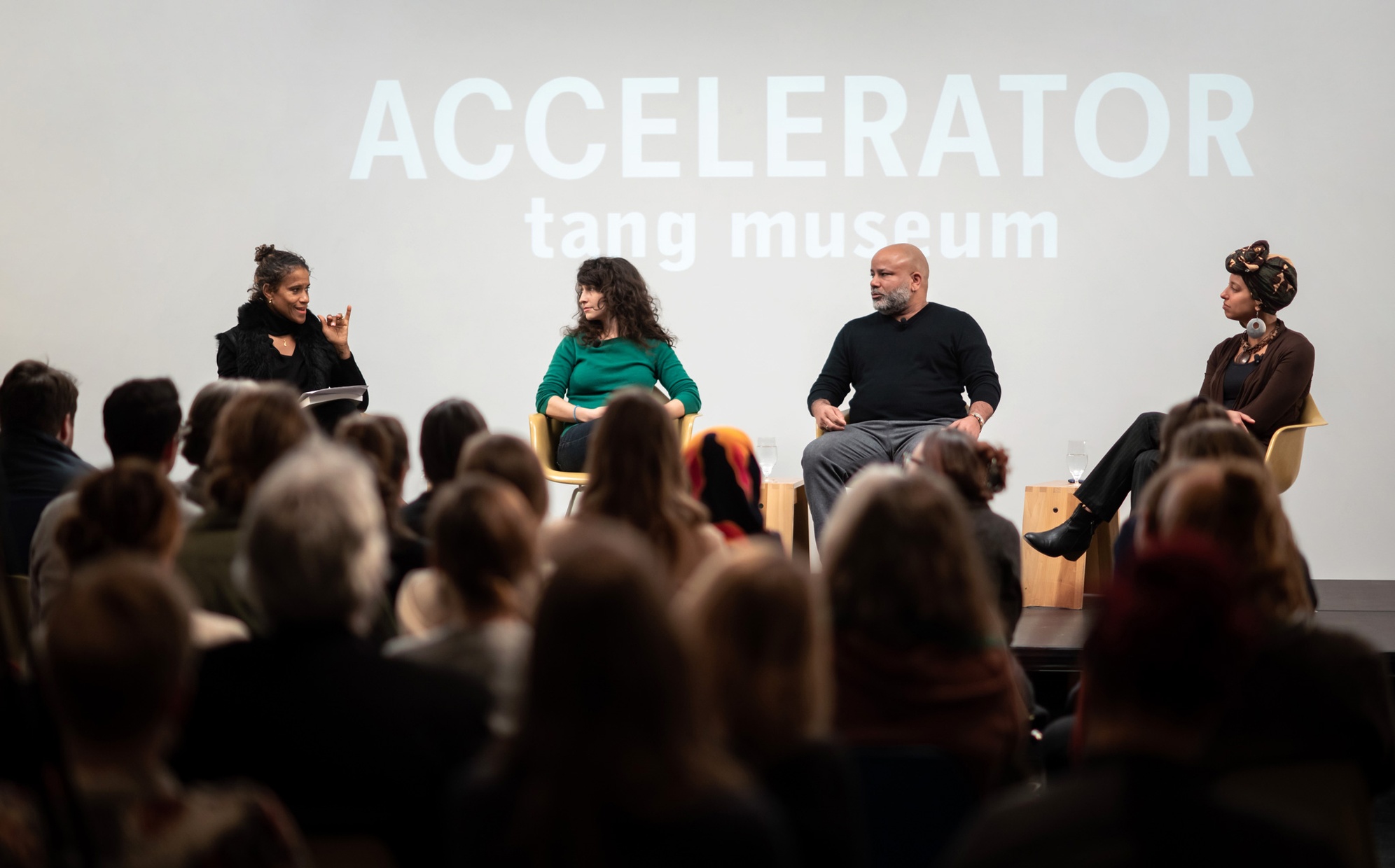 Four people, two black women, one black man, and one white woman, sit on a stage in front of a crowd with the words "ACCELERATOR tang museum" projected behind them.