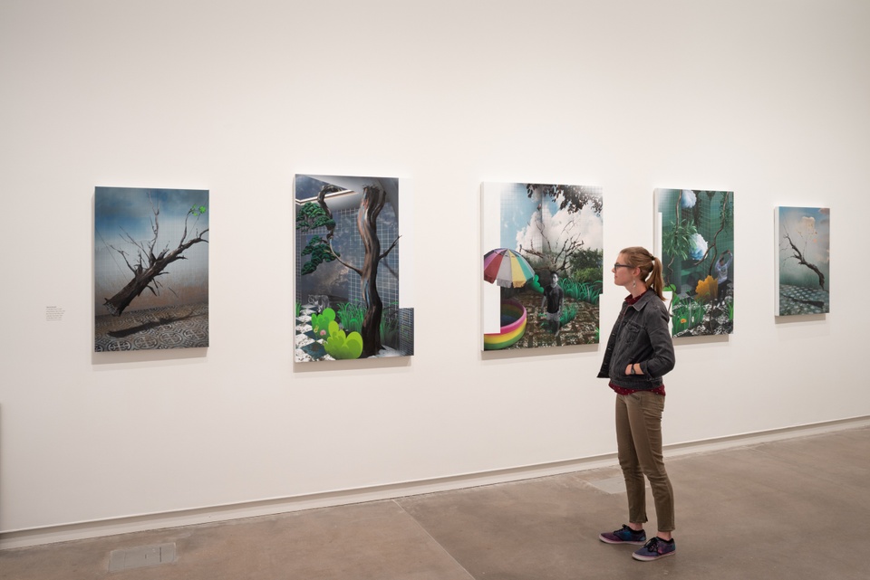 A person views five paintings in a row. The paintings are surreal scenes that have both natural tree and foliage elements and digital elements like grids or pixellated graphics.