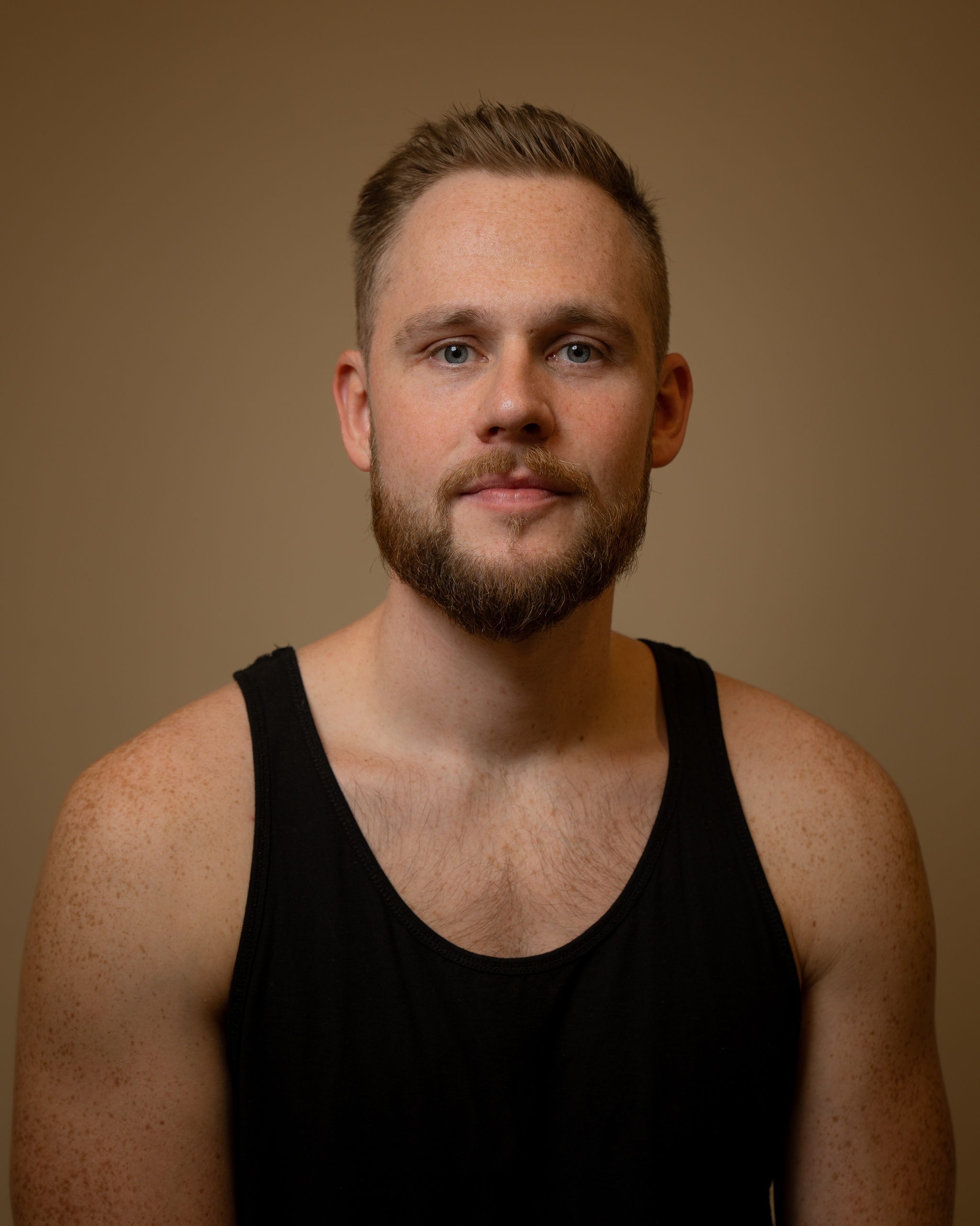 A portrait of Sebastian Charles. Sebastian has short light brown hair and a trimmed bears. He wears a tank top, looking directly at us, while posing against a neutral brown background.