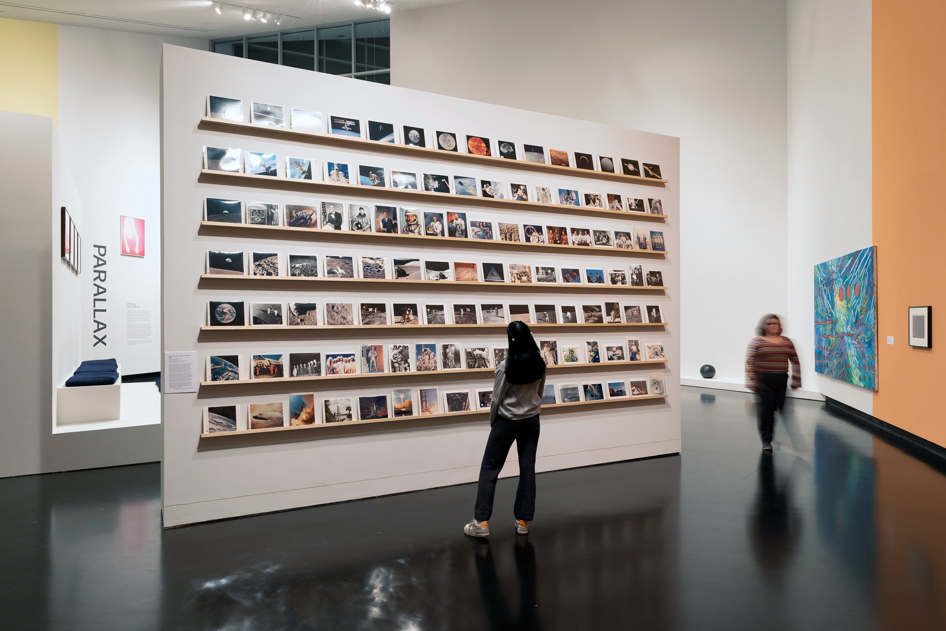 A gallery features a freestanding wall on which sits rows of many small photographs while a young woman with her back to us observes, and another blurred person walks by.