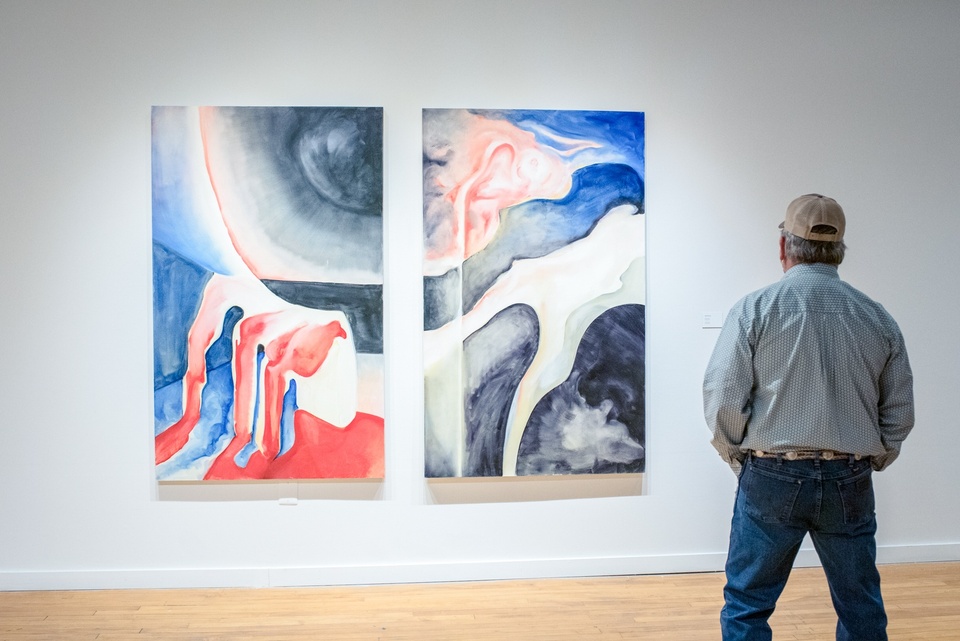 Person stands and observes two paired abstract paintings on a gallery wall, displayed side by side. The paintings together depict a liminal space with some human body elements, done in rich blues, pinks and reds, and grey tones.
