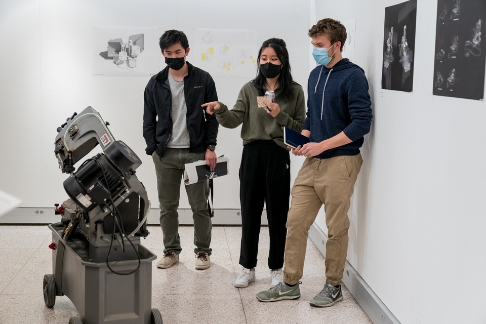 Three people stand in a white-walled gallery space in front of a 3ft-tall wheeled industrial-looking machine. Behind them on the wall are poster prints of digital renderings of machinery.