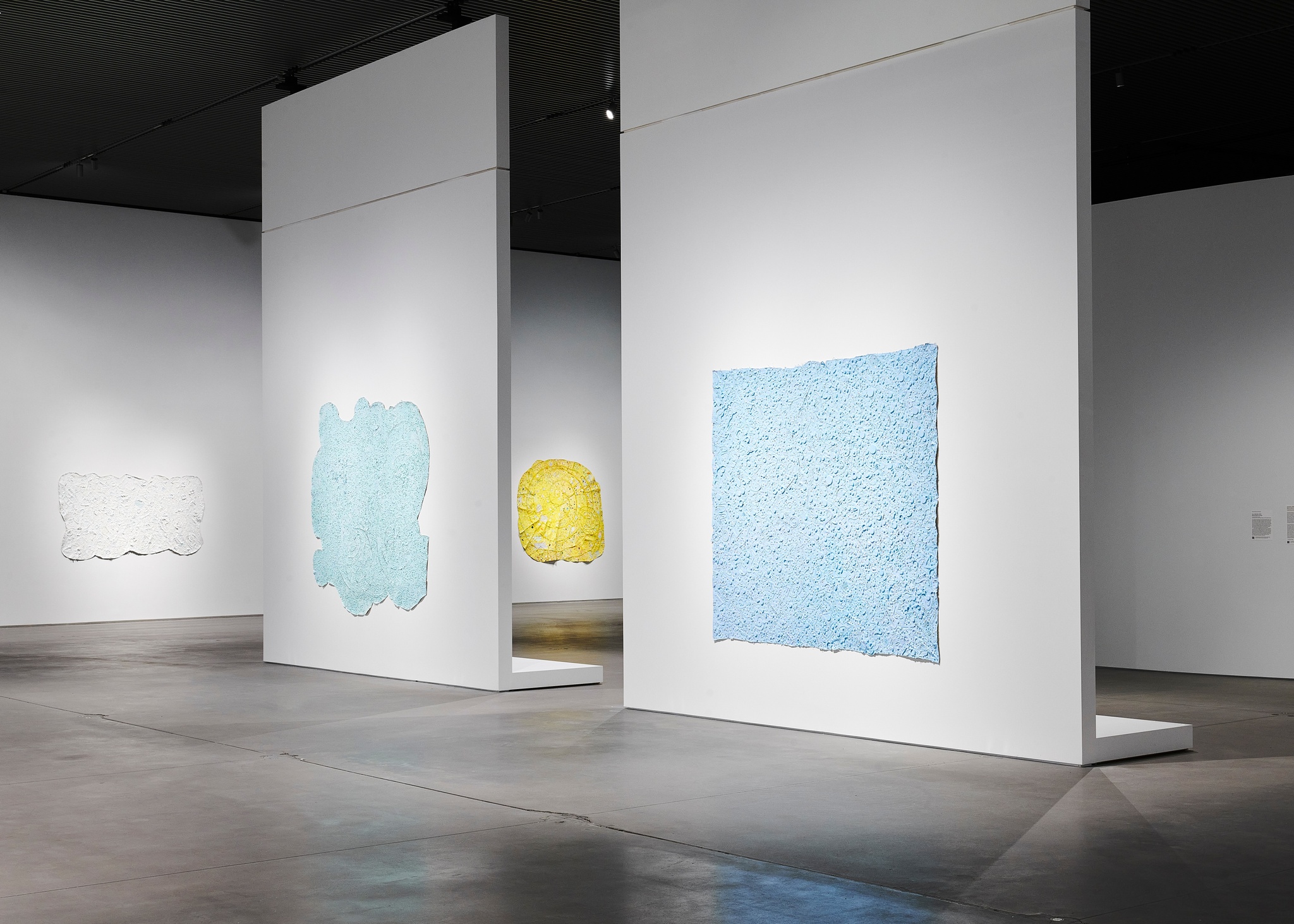 An installation view of abstract paintings in the exhibition Howardena Pindell: Rope/Fire/Water. Two blue and blue-green canvases are on adjacent walls with a curvilinear yellow canvas peeking through a gap in the walls.