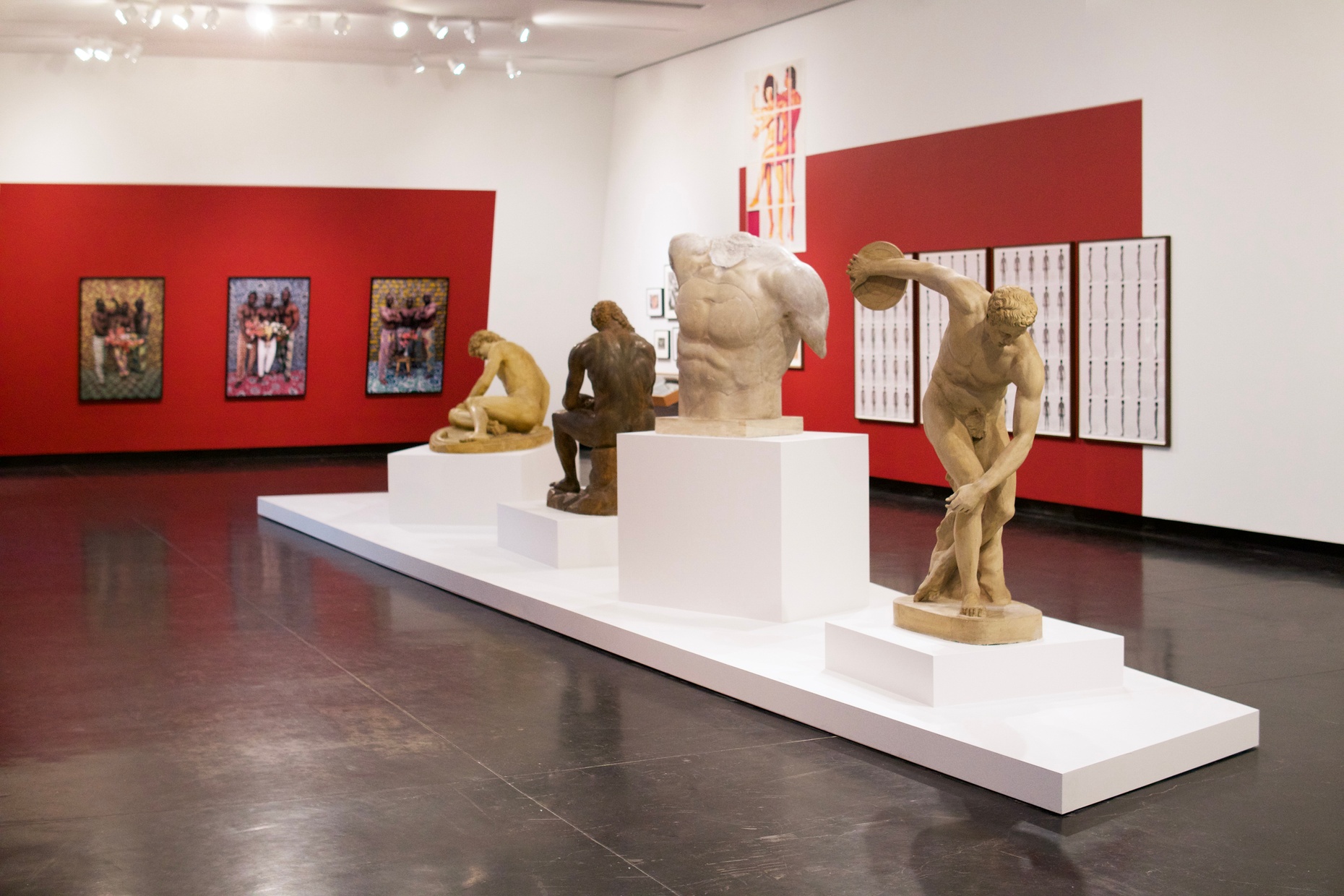 A gallery space with red and white painted walls and artworks featuring muscular people hung up. In the center of the gallery there are four classical sculptures featuring muscular, nude men and a torso.