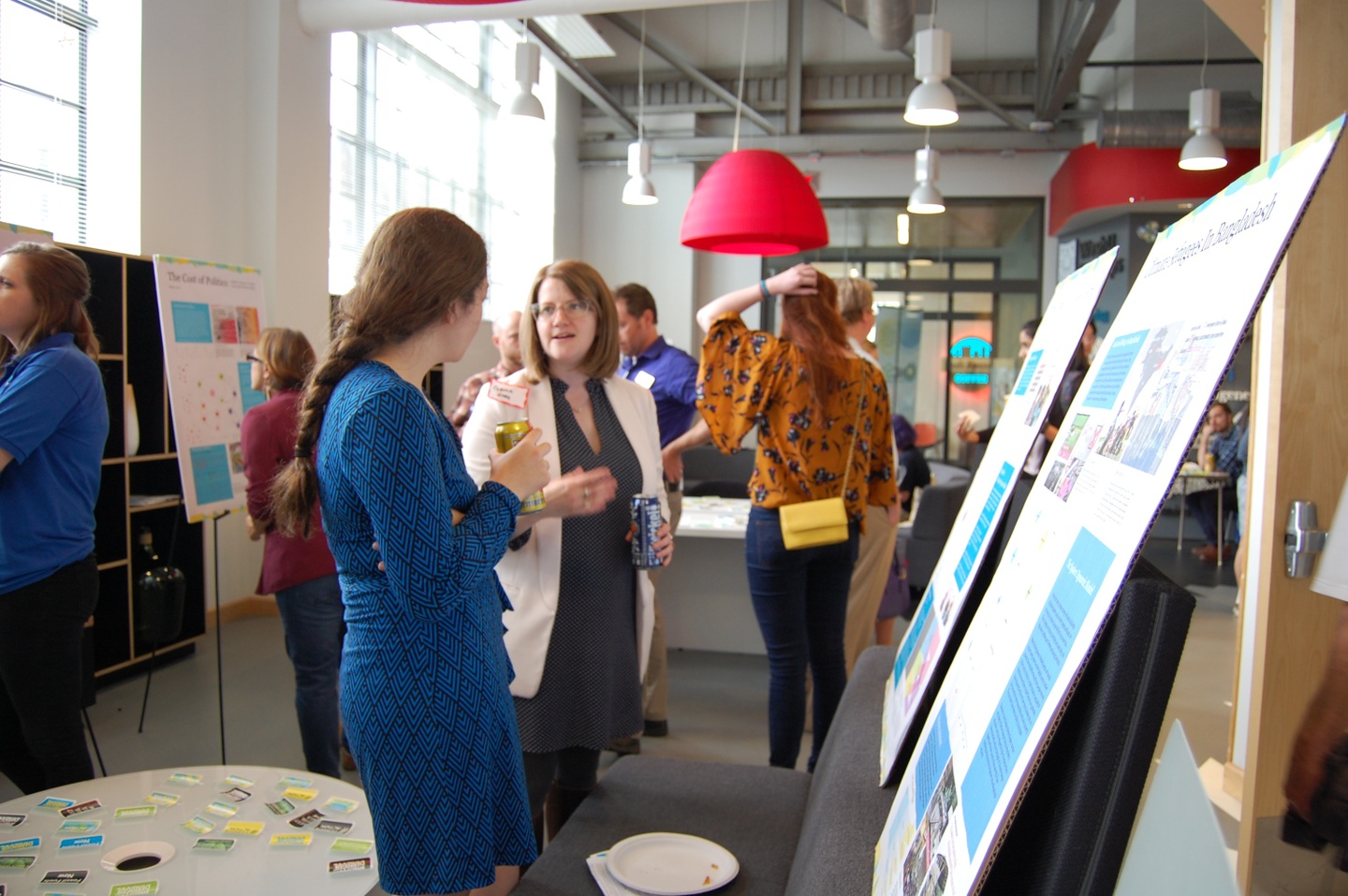 Two people stand in the foreground talking to each other, surrounded by posters and other people.