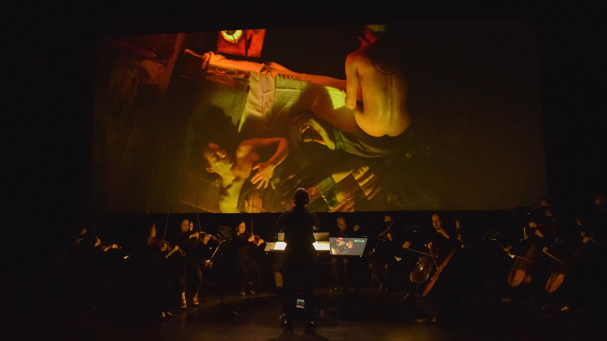 A small orchestra plays in a dark theater space in front of a film screen. On the screen is projected a scene from Wu Tsang's Moby Dick: a naked person sits on the side of a bed while another person still sleeps under a blanket.