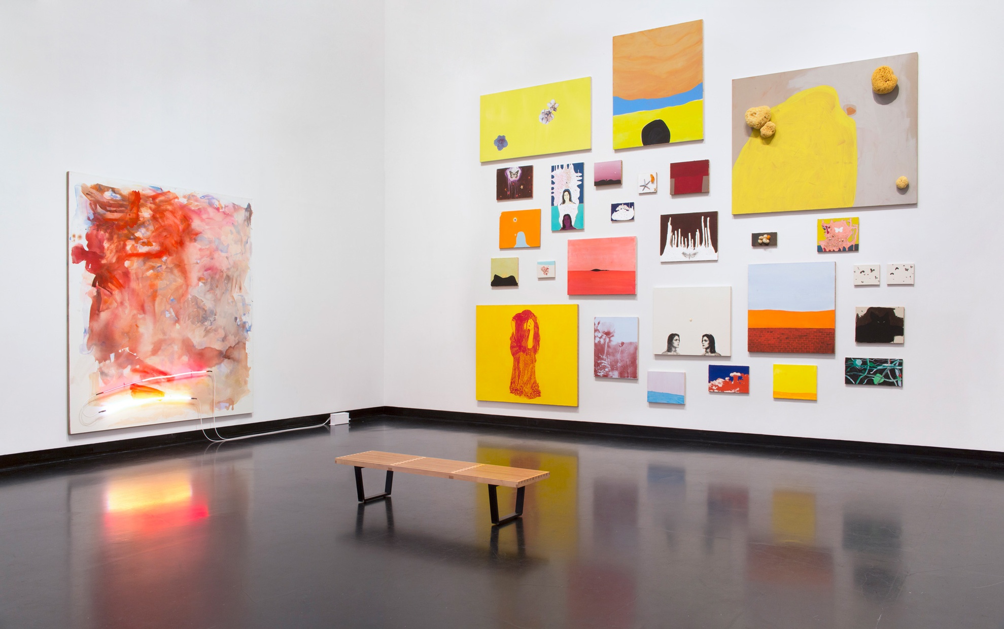 Numerous colorful, abstract paintings arranged on a white wall adjacent to a large, abstract red painting with a yellow neon tube attached near the bottom.