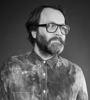 A black and white portait of a white man with a mustache and beard wearing glasses. He looks to the side of us, almost in three quarters profile. He wears a shirt buttoned up to its collar. The shirt has a distressed tie dyed pattern.