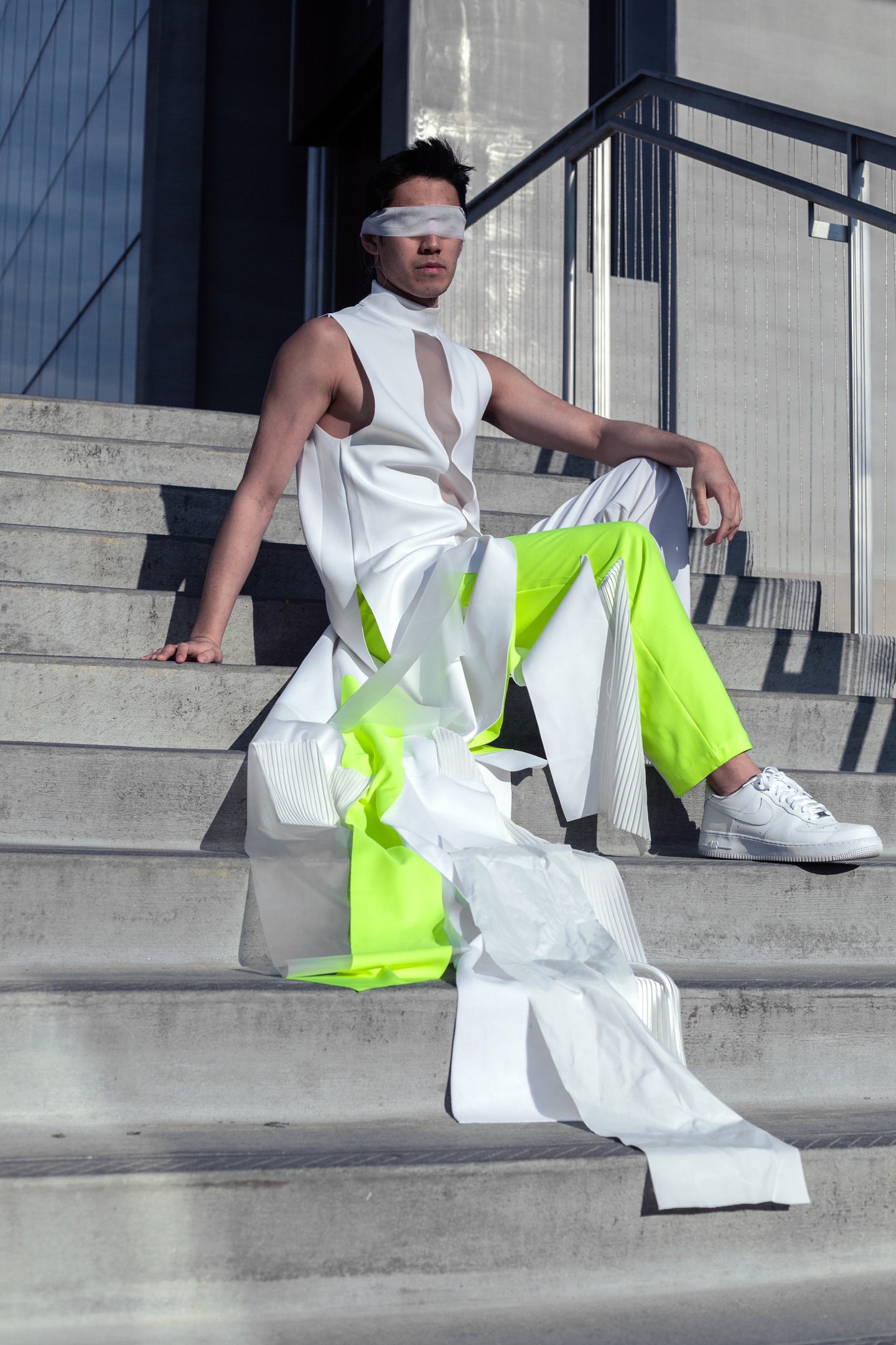 Model sits on concrete steps wearing a high-necked white sleeveless shirt with a see-through gauze panel down the front. The pants are neon green. The shirt and pants both have trailing floor-length strips of fabric in white, gauze, and neon green attached. The model wears a strip of gauze tied across their eyes.