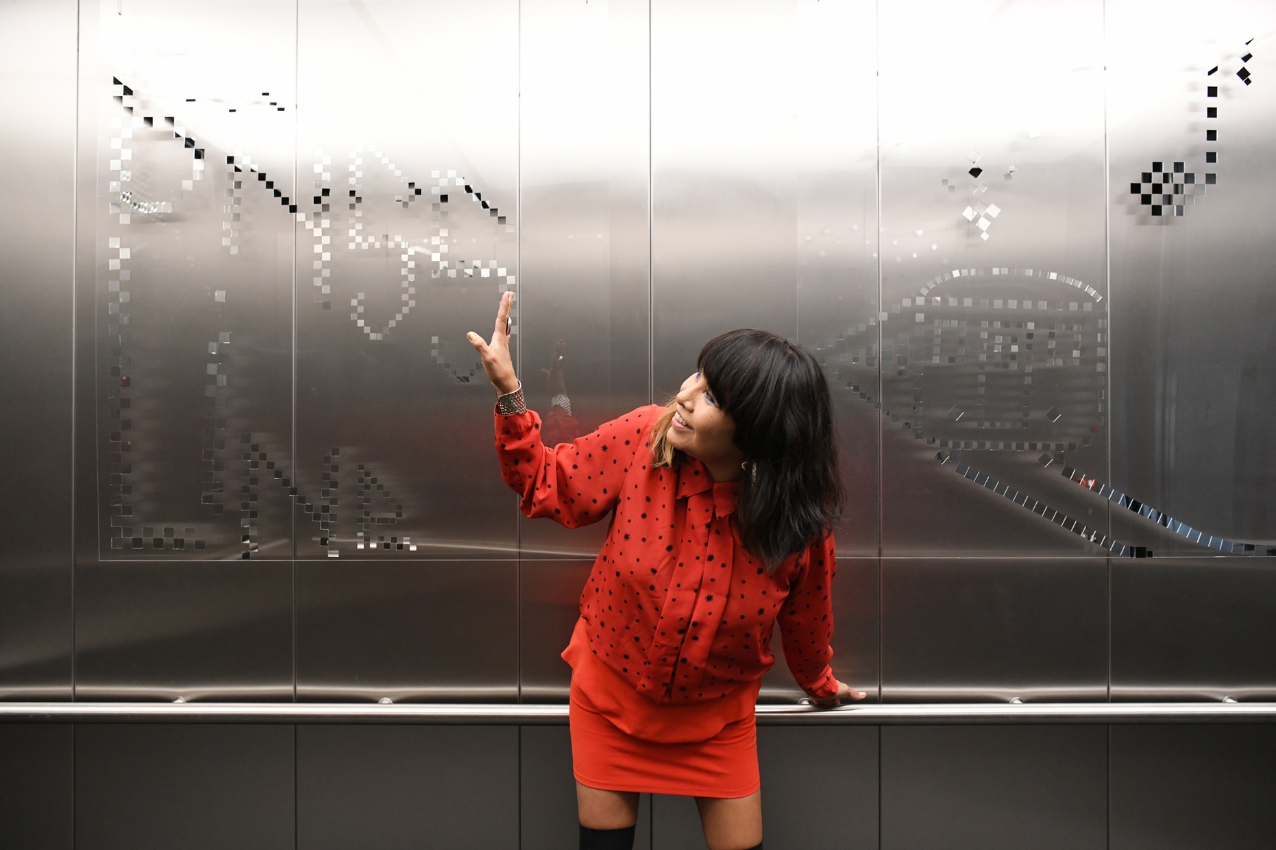 A dark-haired woman wearing a red dress looks and gestures with one arm at a pattern on the wall behind her.