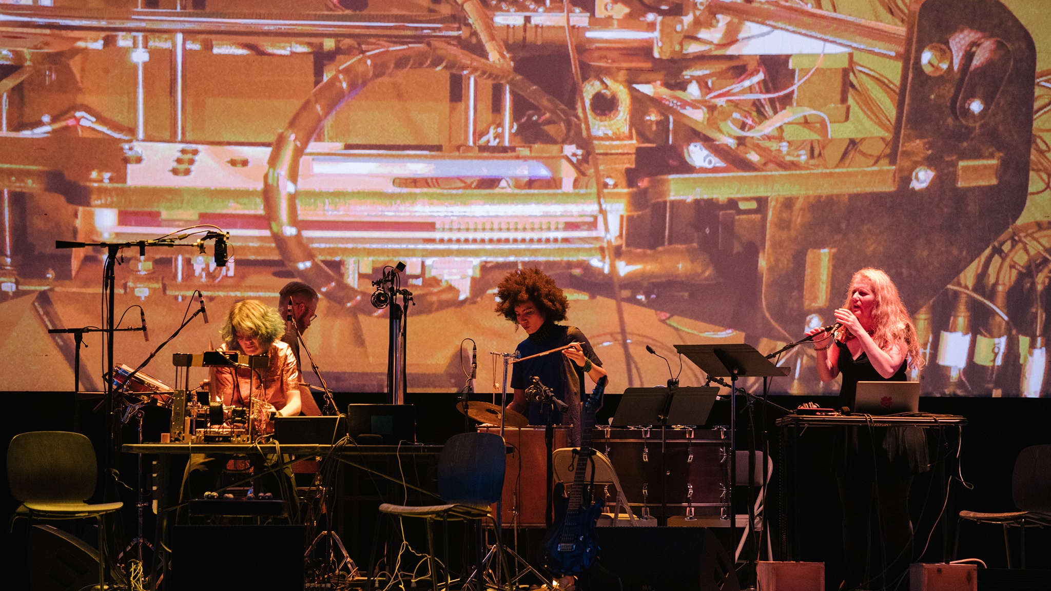 Three musicians on stage perform amid keyboards, microphones, and other instruments. Behind them is a large projection of a close up of technological machinery. 