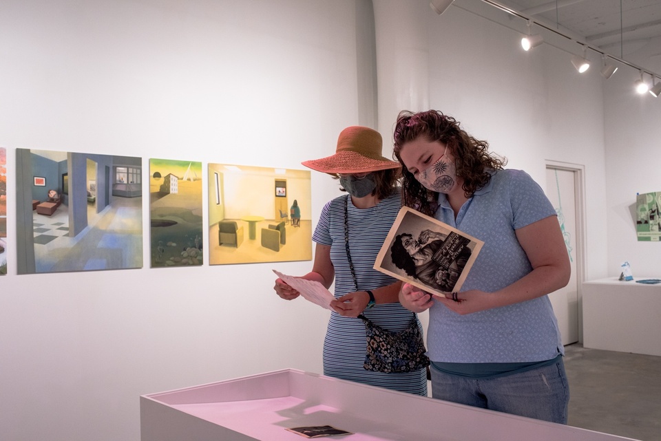 Two people looking at printed pieces from a display in a gallery space exhibiting illustration work.