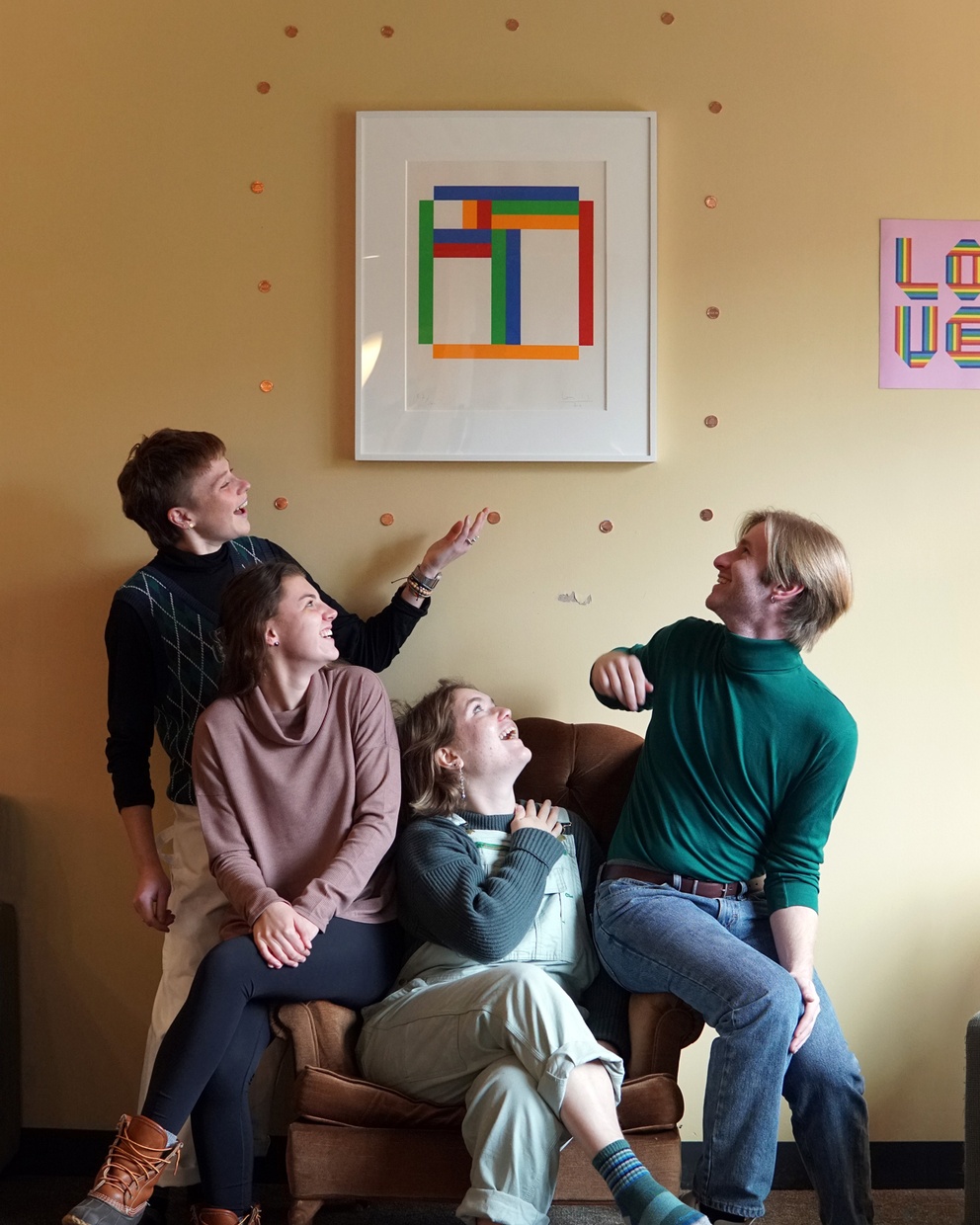 Four white students sit on a plush brown chair and smile as they look up at a work of art hanging on their wall.