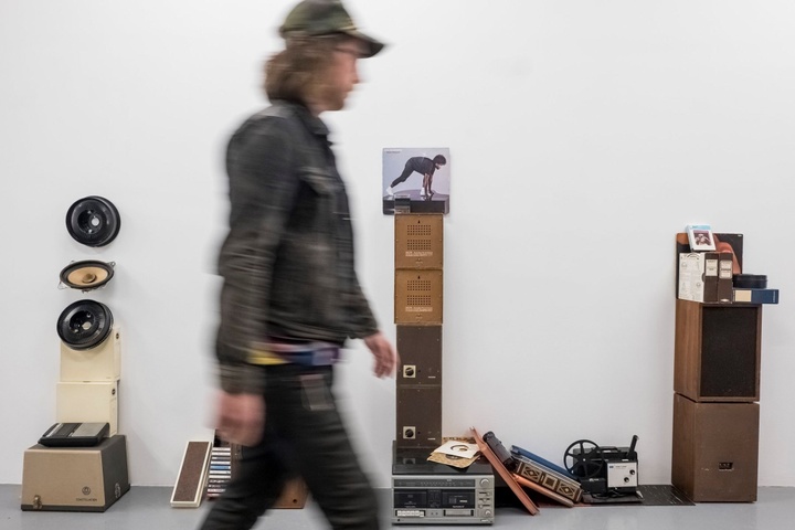 Blurred image of a person walking past three tower-like sculptures installed in a white-box gallery.
