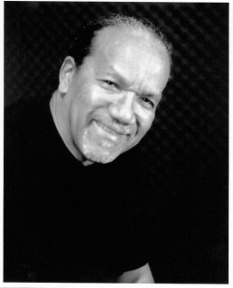 A black-and-white headshot of dancer and founding artistic director Rod Rodgers. A Black man wearing a black t-shirt, he leans slightly in the image with his head turned as he smiles warmly.