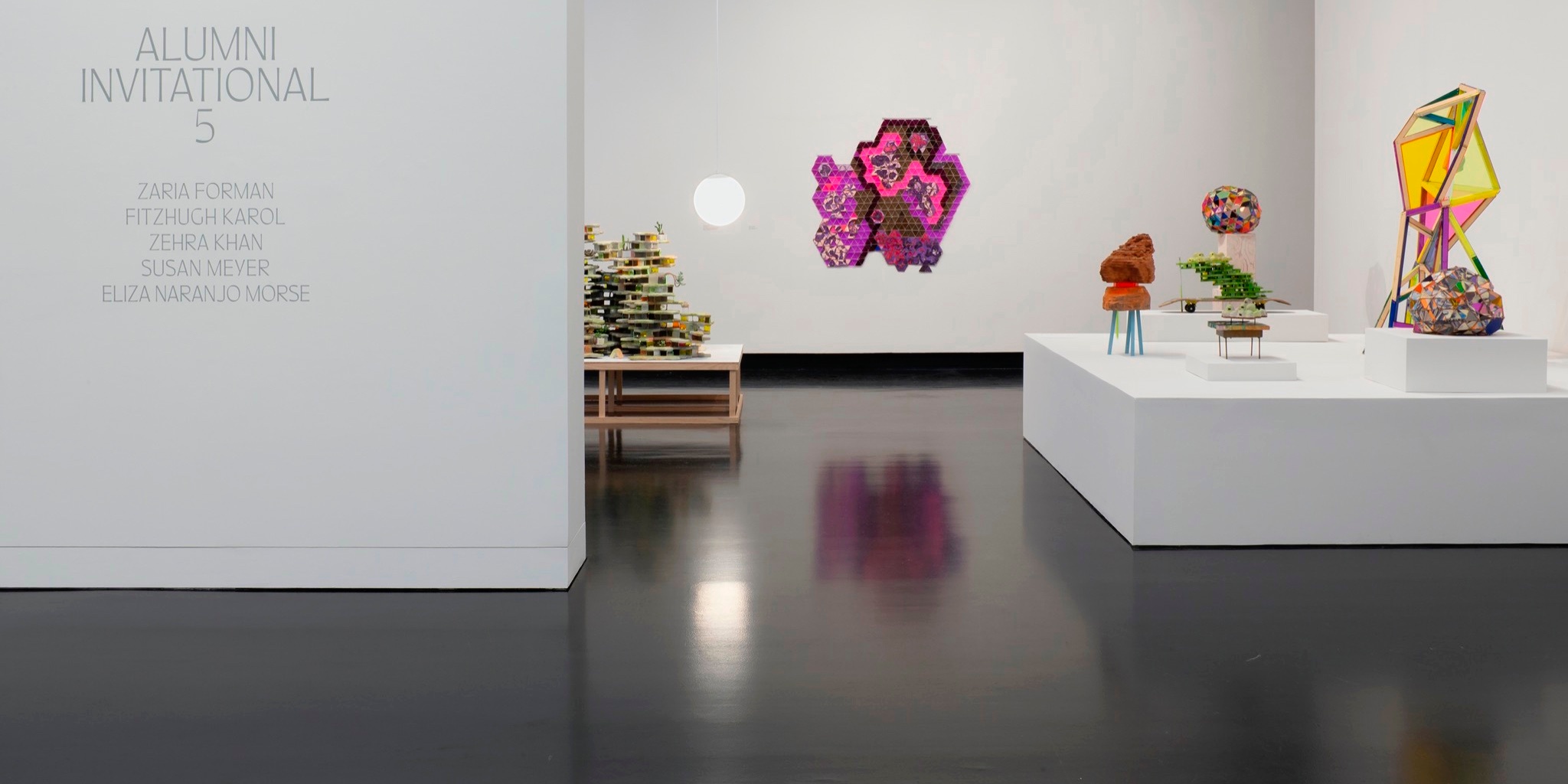An interior of a museum gallery with one wall showing the title Alumni Invitational 5, with a number of colorful sculptures sitting on various plinths, and hung on the wall.
