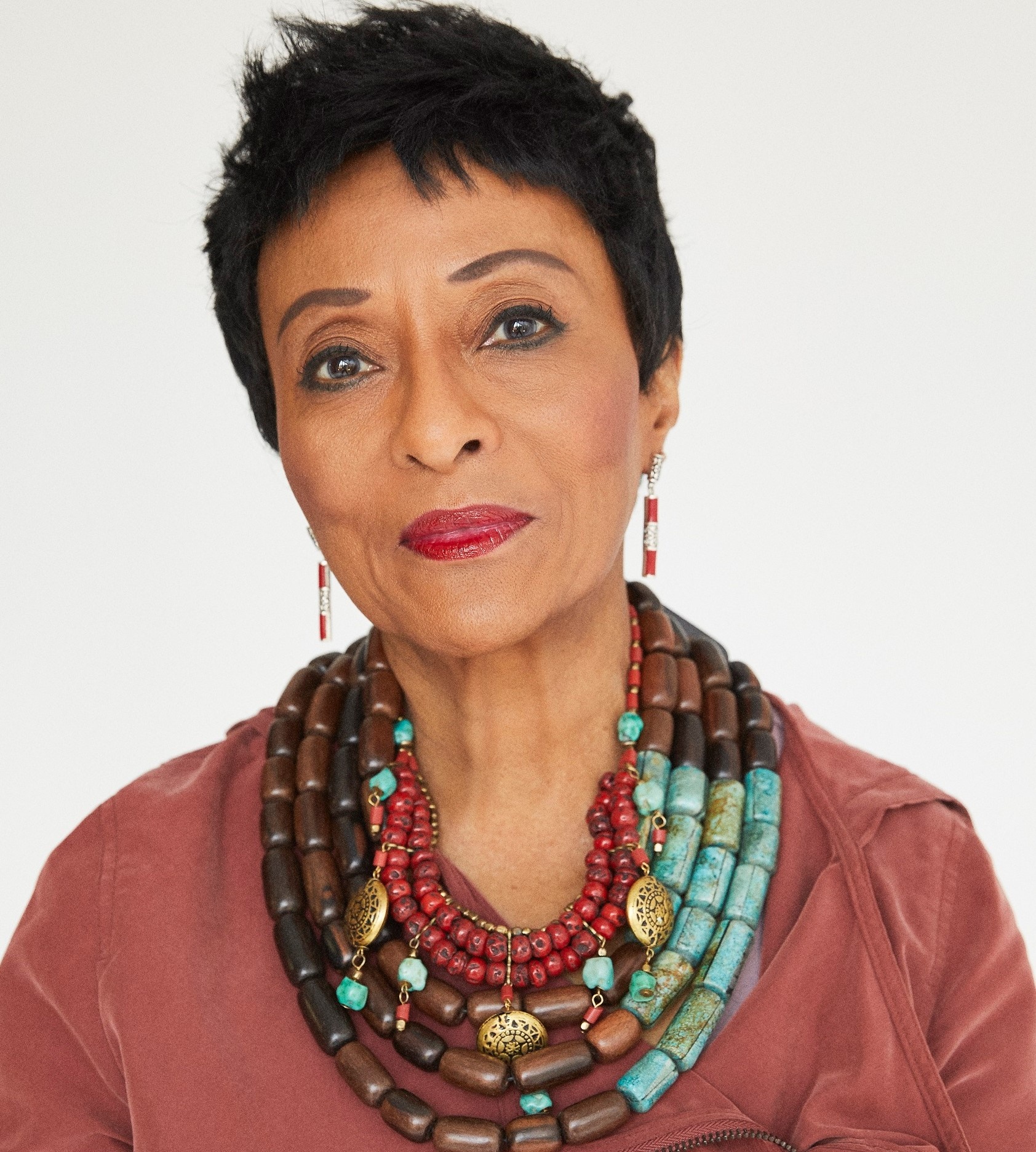 A Black woman with short-cropped dark hair arches one eyebrow. She wears bright red lipstick and a necklace with large brown and turquoise beads looped around her neck several times.  