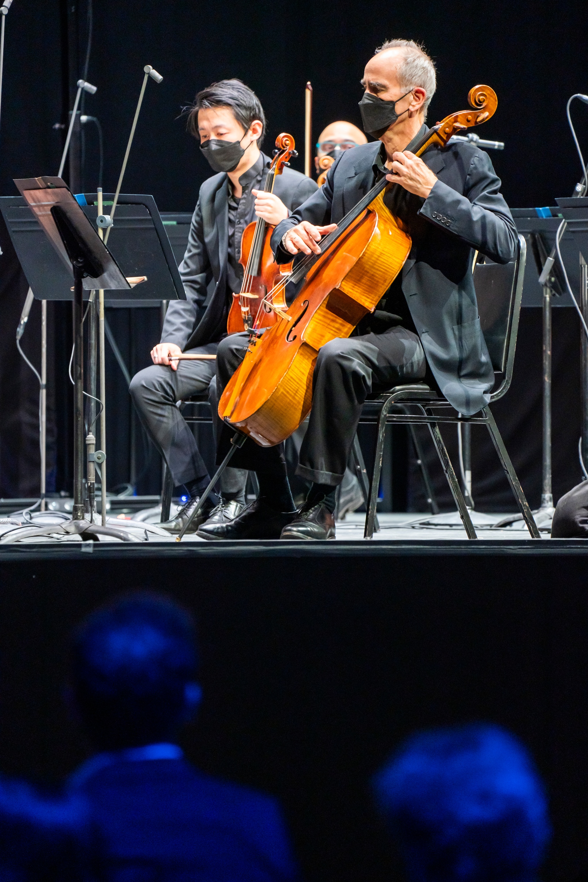 A cellist and violist seen on stage with an audience sitting in shadowy blue light beneath and in front of them.