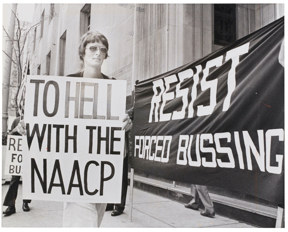 A black and white photograph of a woman wearing sunglasses holding a handwritten sign that reads “TO HELL WITH THE NAACP” with a large building and other sings and people in the background.
