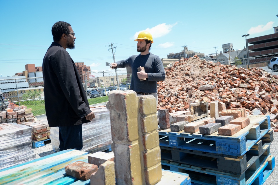 Two people talk in front of a pile of bricks, with a stack of gold bricks in front. 