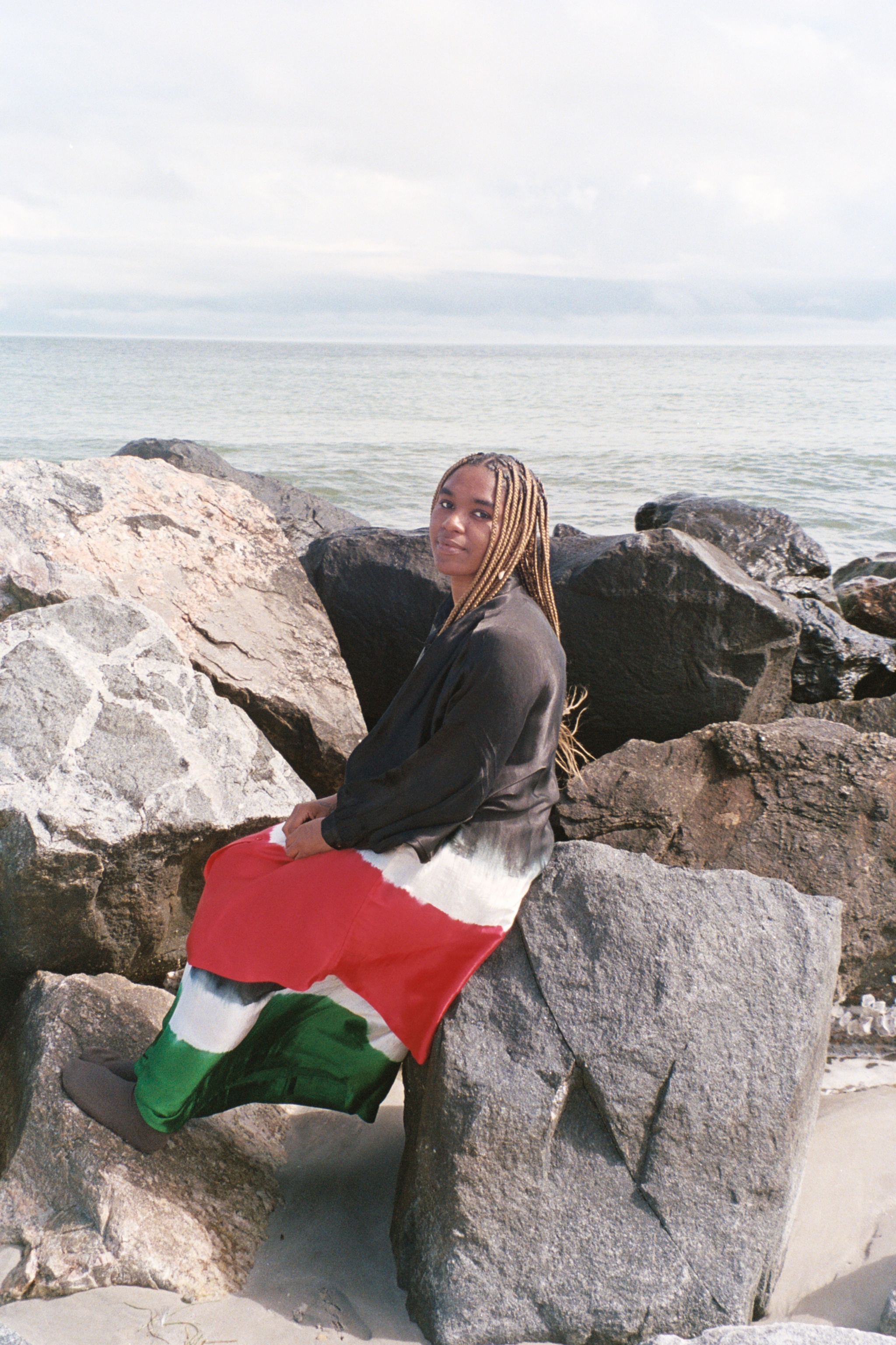Yaz Lancaster, a nonbinary Black artist, sits on large boulders along a seashore. They have long locs that hang beneath their shoulders. Yaz looks back at us over one shoulder. They wear a long dress with bands of color: black, white, red, and green.