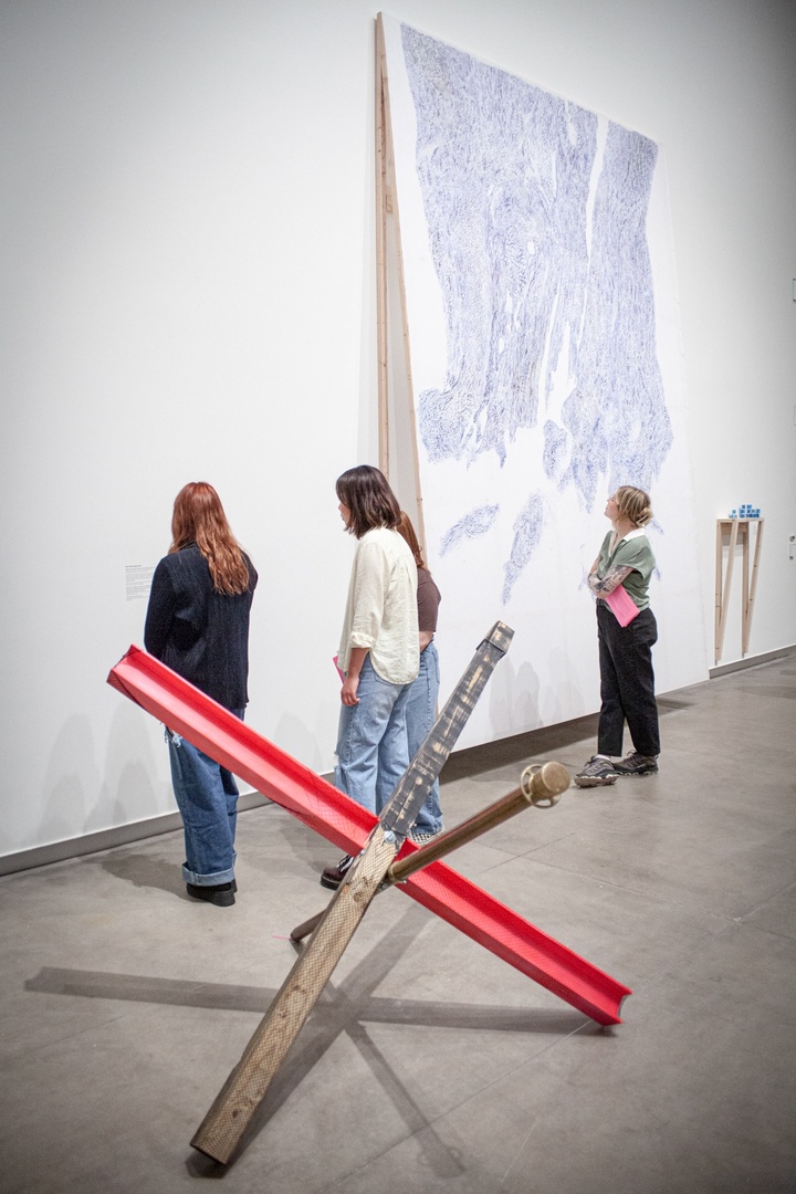 A sculpture in the shape of a jack sits on the floor of the gallery. The three arms of the jack shape are made of a charred piece of lumber, a brass stanchion, and an I-beam painting hot pink. The legs of the structure are encased in fishnet stockings.