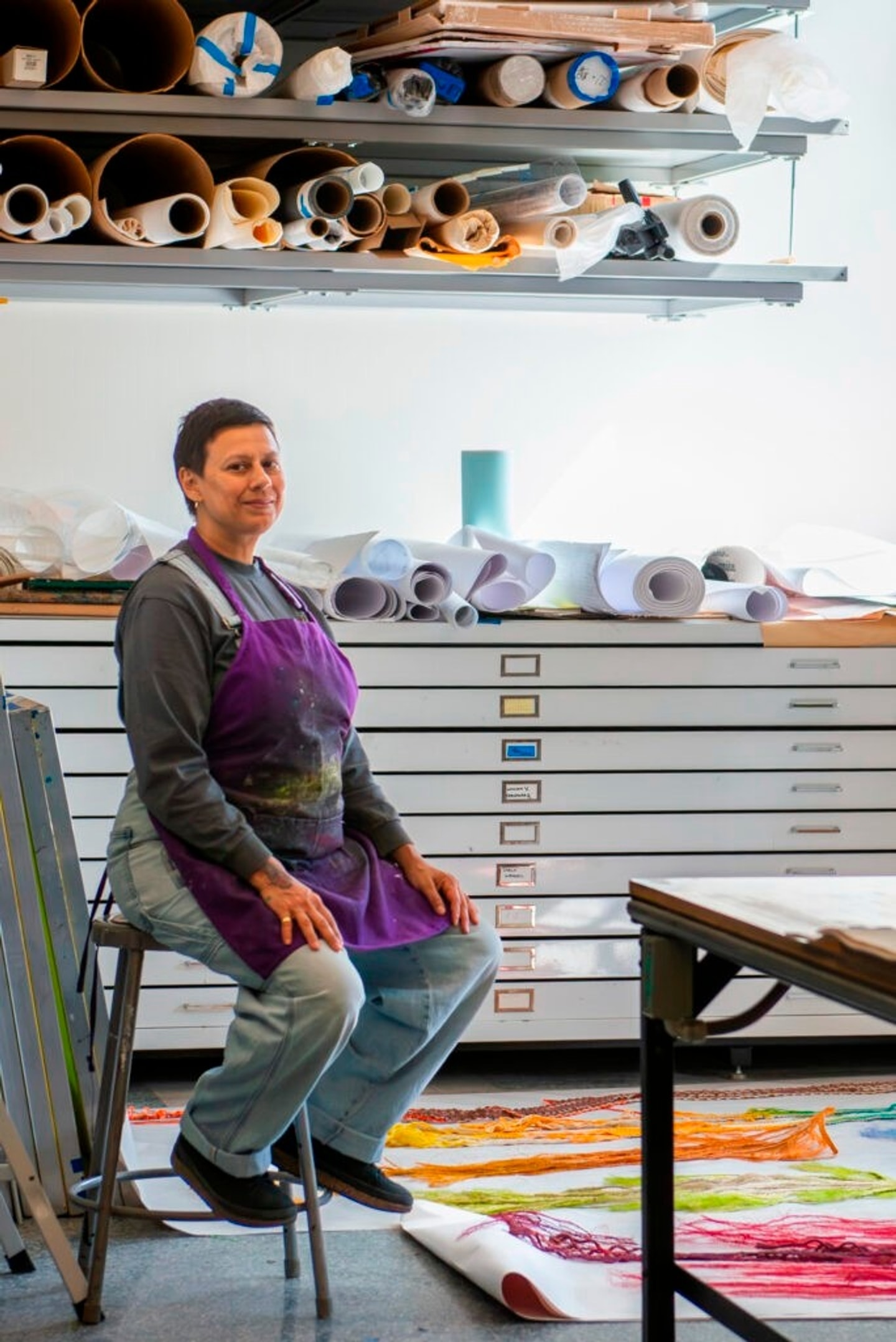 portrait of Carolina Caycedo in a printmaking studio sitting on a stool and looking at the camera 